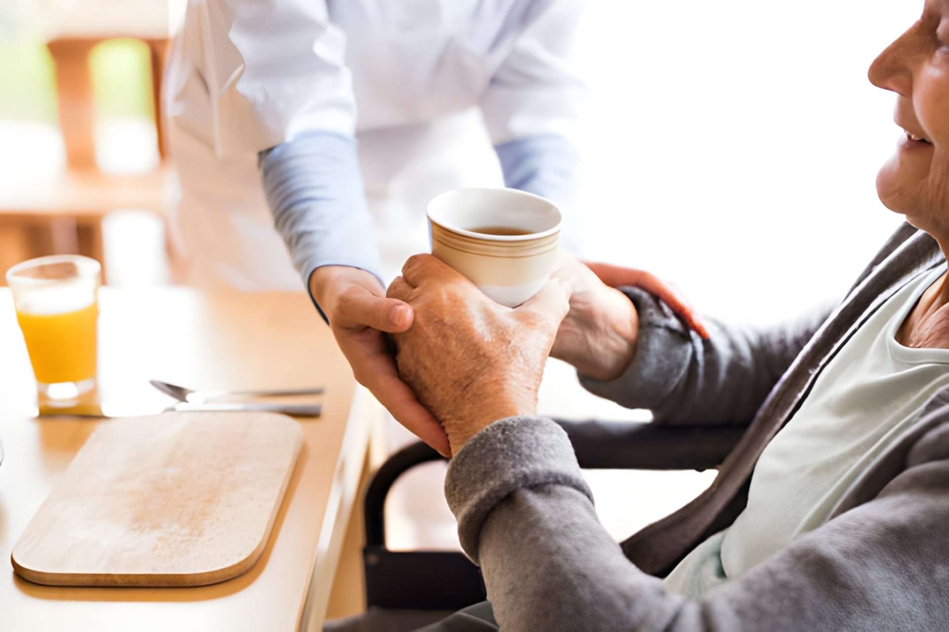 A nurse giving tea to an elderly woman sitting at the table. 